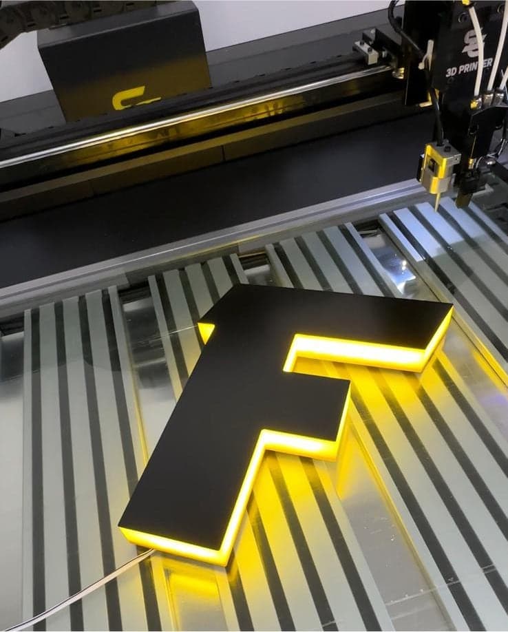 A letter F with a black face and a yellow halo glow, on the printer bed under the print head