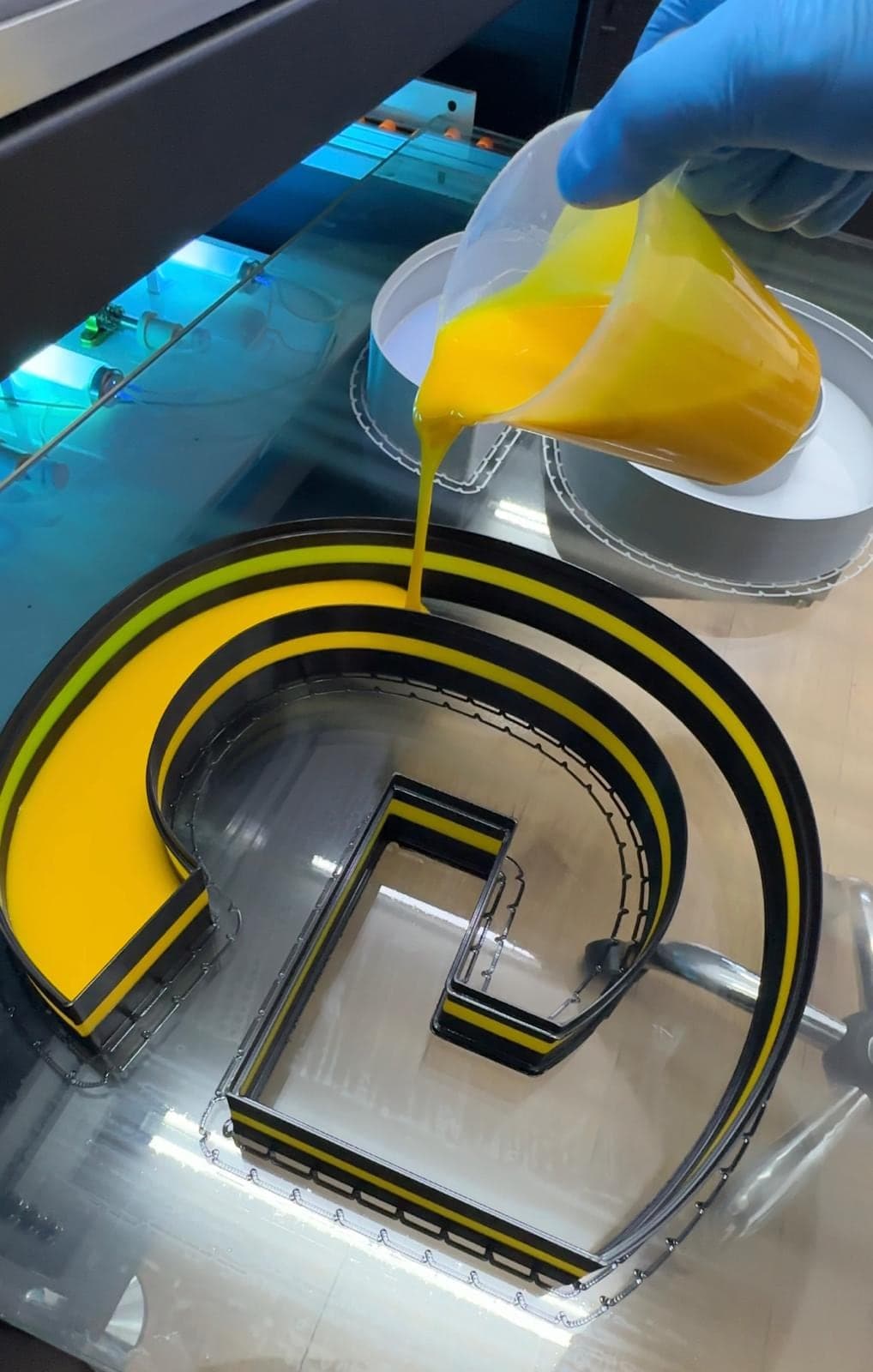 A gloved hand pouring yellow liquid acrylic into a printed letter G lying face-down on the bed