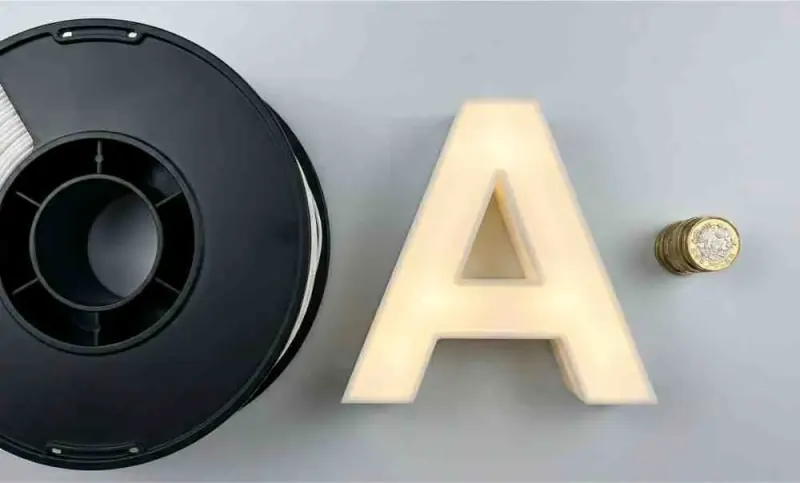A filament spool, a finished 3D-printed letter and a few pound coins, showing the low material cost per letter
